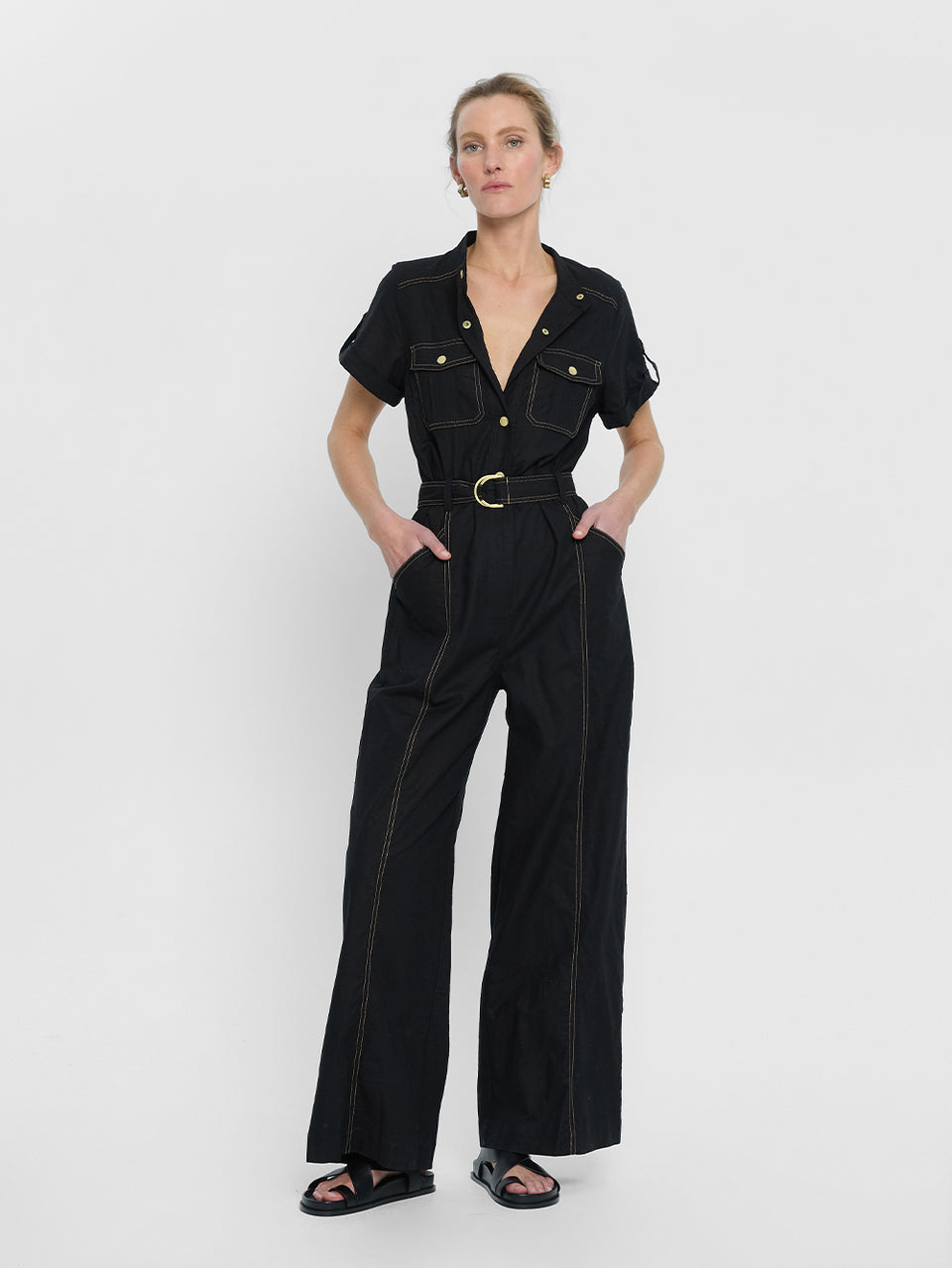 tudio model wears the KIVARI Ebony Jumpsuit: a black linen jumpsuit with gold buttons, topstitch details, a button-front with signature hardware and belt.