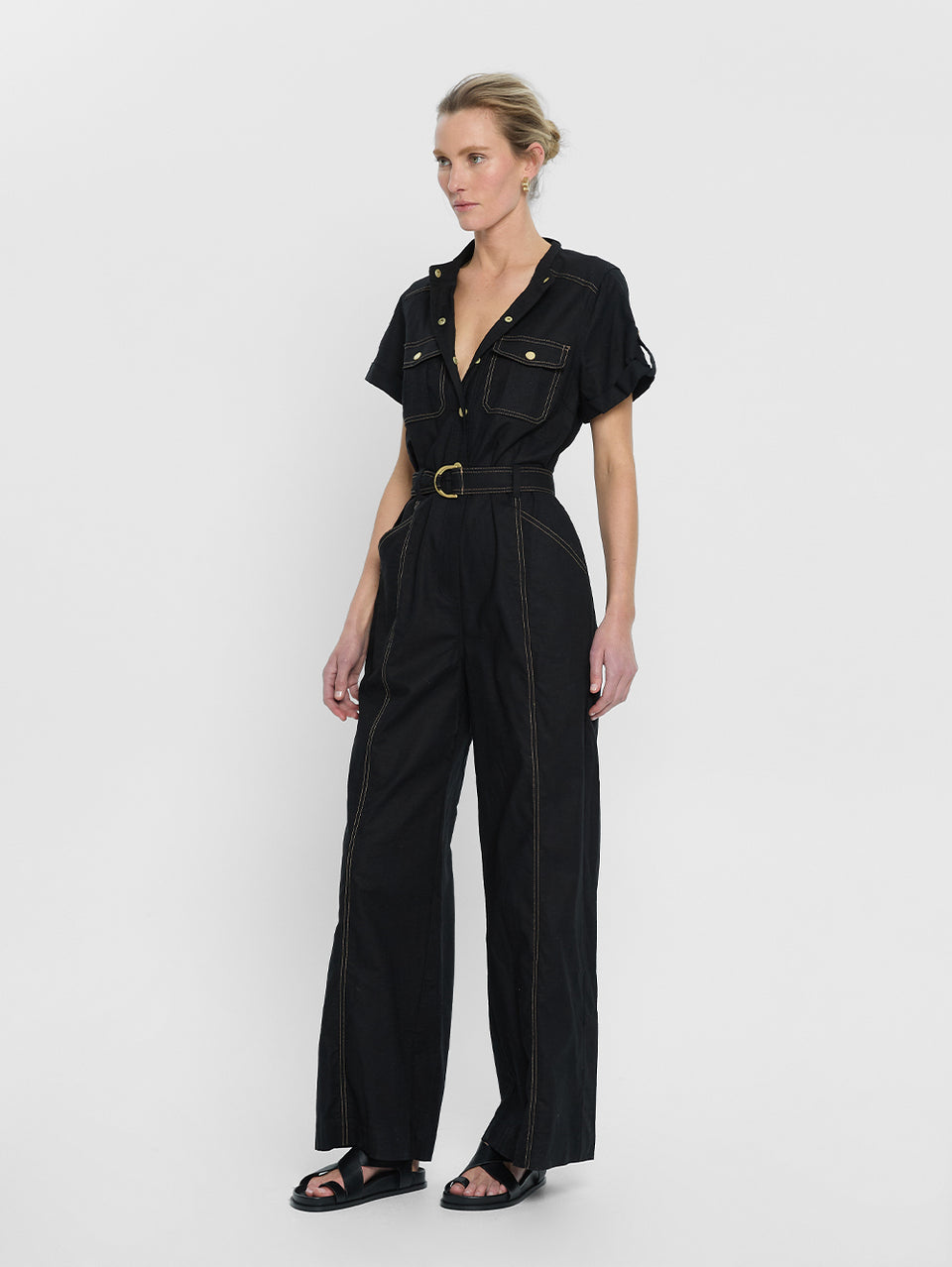 tudio model wears the KIVARI Ebony Jumpsuit: a black linen jumpsuit with gold buttons, topstitch details, a button-front with signature hardware and belt.