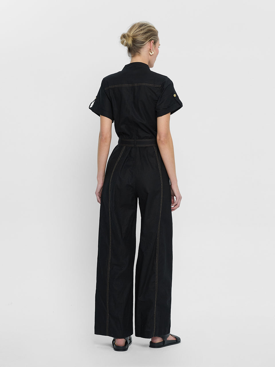 tudio model wears the KIVARI Ebony Jumpsuit: a black linen jumpsuit with gold buttons, topstitch details, a button-front with signature hardware and belt.