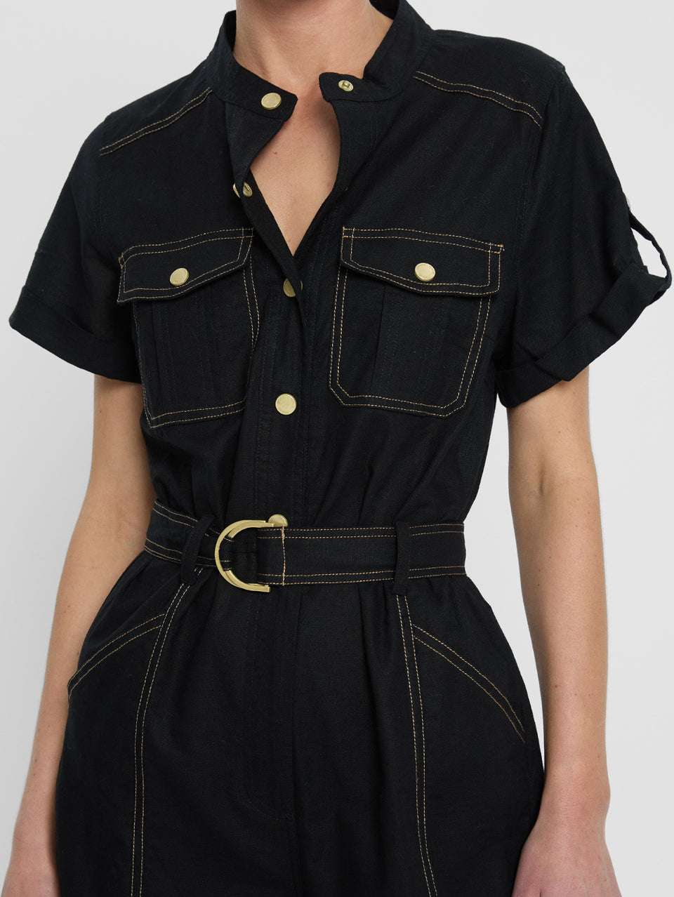 tudio model wears the KIVARI Ebony Jumpsuit: a black linen jumpsuit with gold buttons, topstitch details, a button-front with signature hardware and belt.