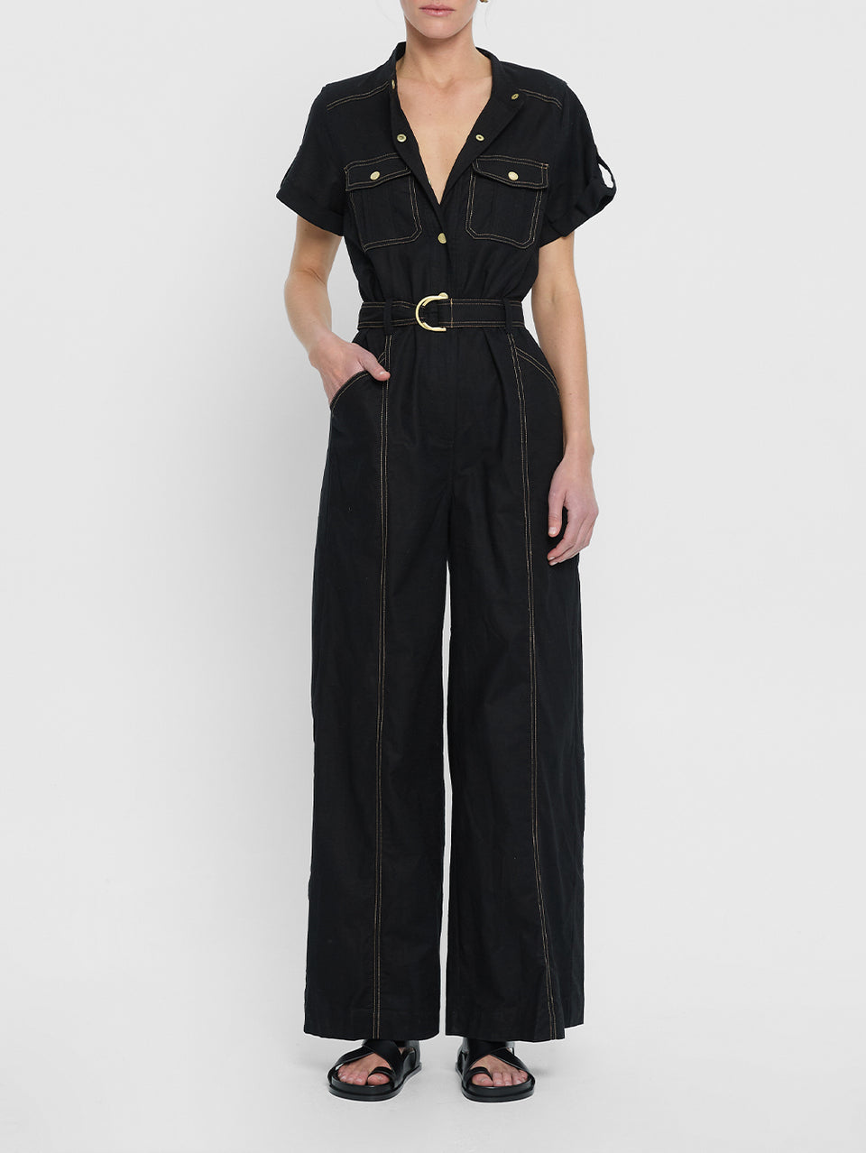 tudio model wears the KIVARI Ebony Jumpsuit: a black linen jumpsuit with gold buttons, topstitch details, a button-front with signature hardware and belt.